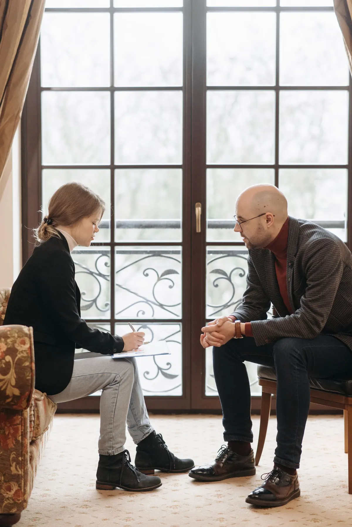 Two people sitting facing each other by a tall window, one taking notes.