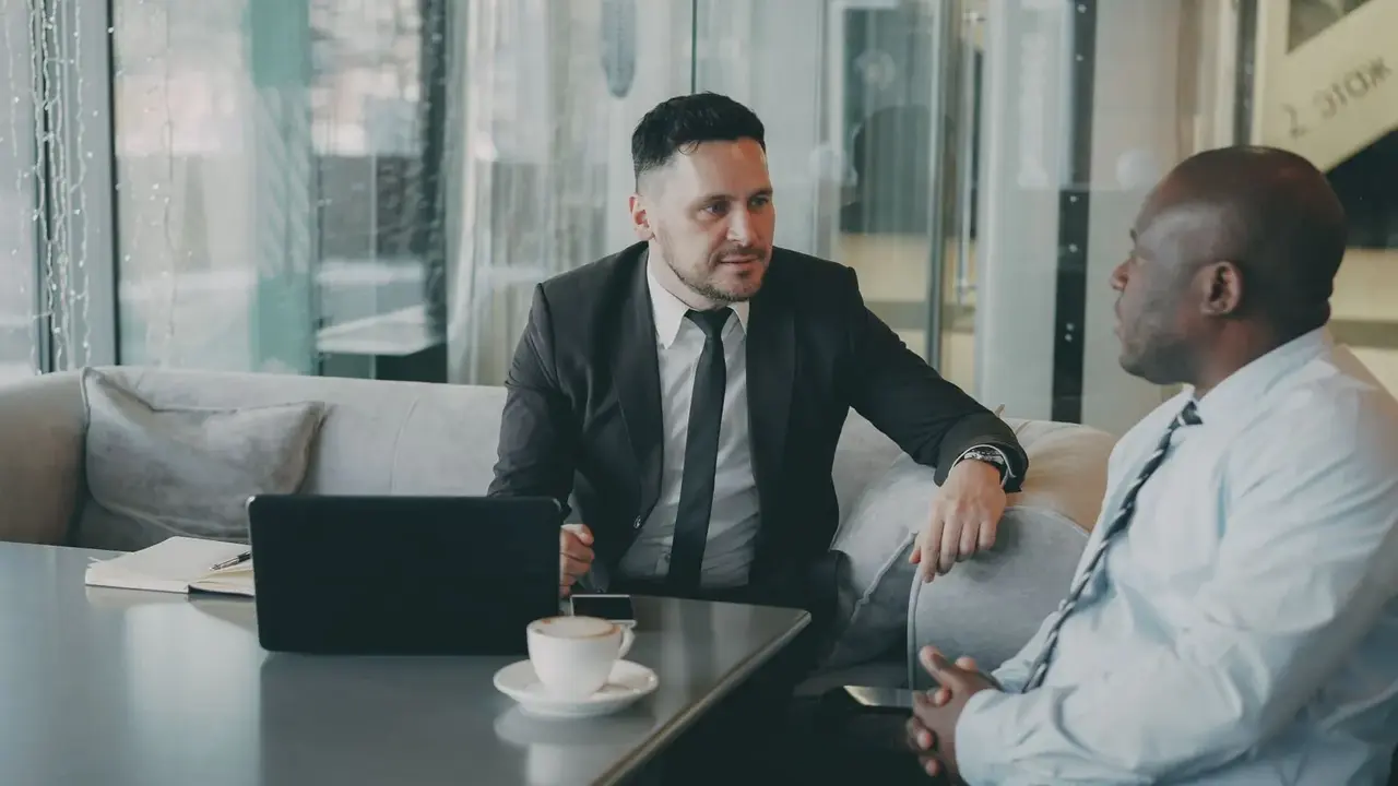 Two men in suits talking across a low table with coffee between them.